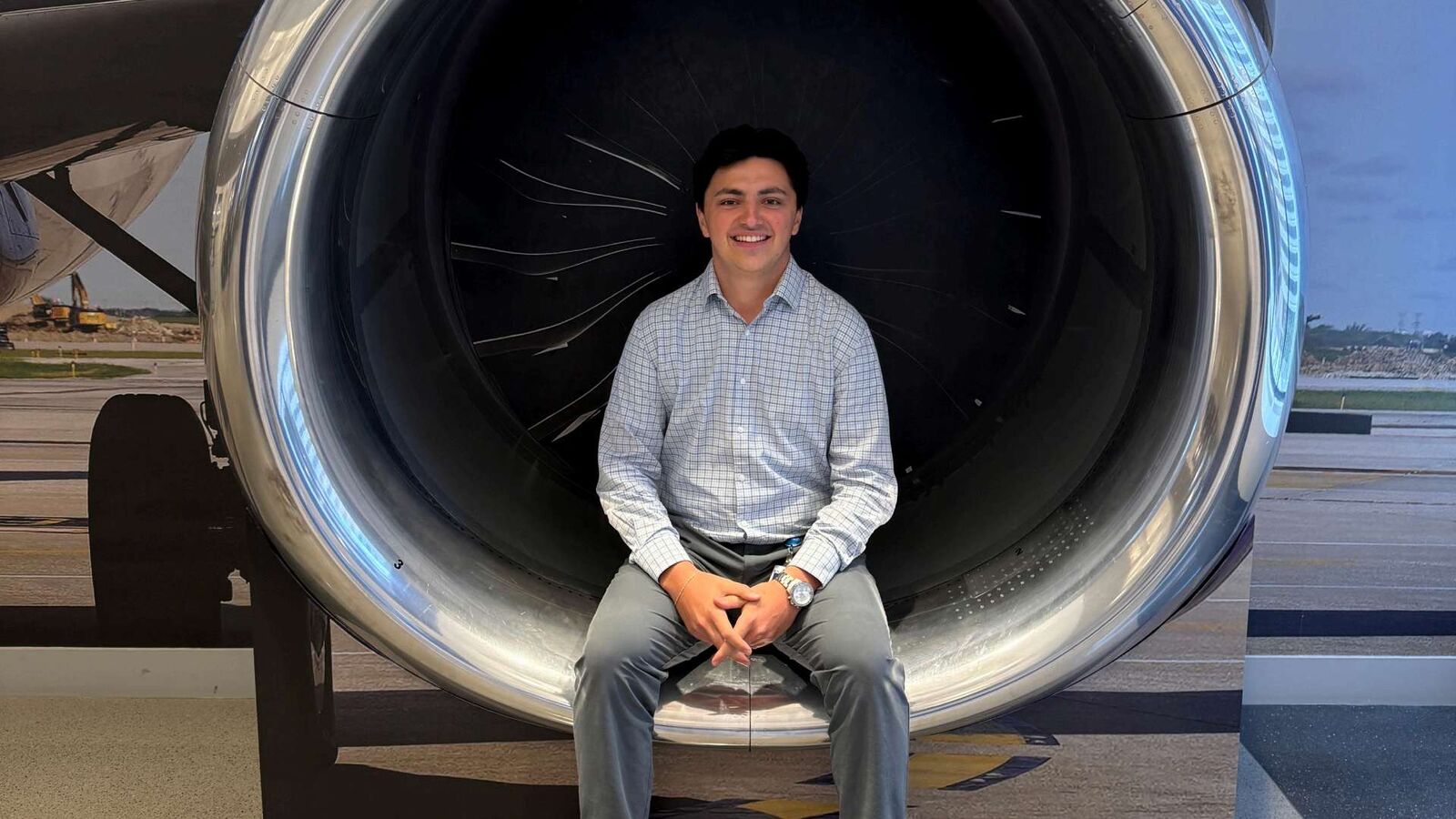 Sam Rizzo seated in front of a jet engine display, wearing a checked shirt.